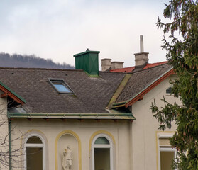 Roof complex of an old building with decorative elements, chimneys, and a green extension in an urban environment