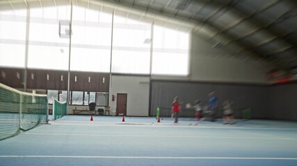 empty nearcourt foreground with scattered tennis ball, cones and distant players running group drills, blurred motion and wide perspective, coaching station visible near wall, communal training vibe