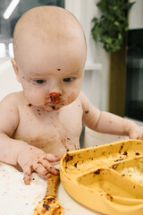 Toddler covered in food sits in a high chair and enthusiastically eating for meal. Baby playfully messy eating. Concept of early childhood nutrition and introducing solid foods to infants.