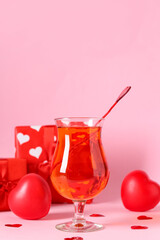 Glass of tasty cocktail with hearts and presents on pink background, closeup. Valentine's Day celebration