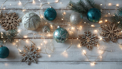 Sparkling silver and teal Christmas ornaments, wooden snowflakes, pine branches, and twinkling fairy lights arranged on a rustic white wooden background.