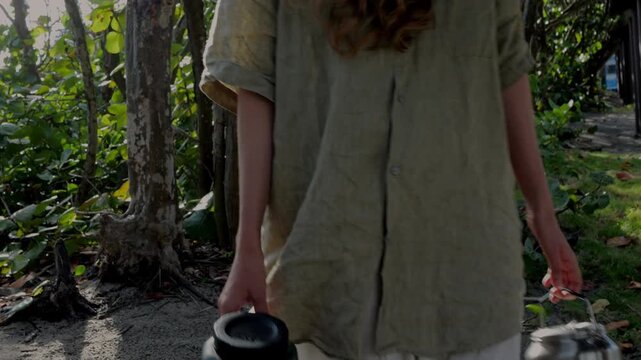 Woman walks through coastal forest carrying water container and metal camping kettle. Concept of camping, camp chores and simple outdoor living.