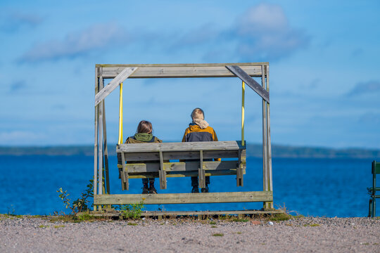 Children seated on rustic wooden swing facing peaceful ocean horizon on clear day.