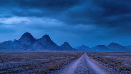 Fototapeta premium A long, solitary gravel road extends through a vast, arid landscape towards majestic blue mountains under a dramatic, cloudy twilight.
