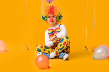 Sad little boy in clown costume with balloons sitting on yellow background. April Fools Day celebration