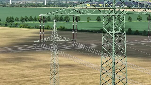 Telephoto drone view of intersecting high-voltage lines and close-up of insulators on a pylon across flat fields, with wind turbines in Saxony-Anhalt, Germany.