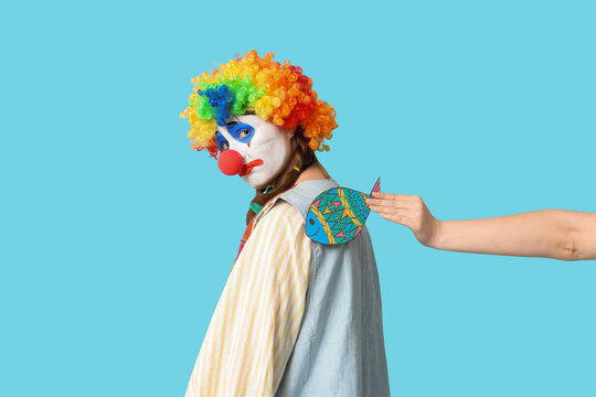 Young woman dressed as sad clown with paper fish attached to her back on blue background. April Fool's day celebration