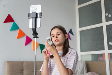 Girl recording a creative vlog at home, showing craft materials in front of a smartphone on a tripod. Young content creator demonstrating glitter supplies in a bright and cozy room.