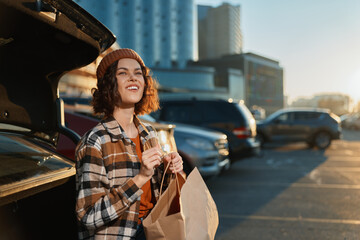 Fototapeta premium Woman by car in parking lot holding a paper shopping bag, smile and sunlight at golden hour glow. Candid lifestyle portrait with authenticity, mindful living and emotional storytelling vibe.
