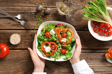 Female hands with bowl of fresh vegetable salad with bell pepper and feta cheese on wooden background