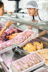 Positive young salesman standing behind counter demonstrating sausages in plastic container in butcher shop