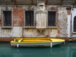 An intimate and evocative view of the Rio de Ca' Widmann in Venice, Italy. A bright yellow and grey-hulled boat is moored alongside a facade of historic bricks and aged plaster.