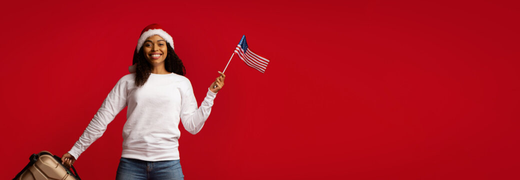 A woman wears a Christmas hat and a casual white shirt while holding an American flag. She stands with a suitcase, smiling happily, in front of a bright red backdrop during the holiday season.