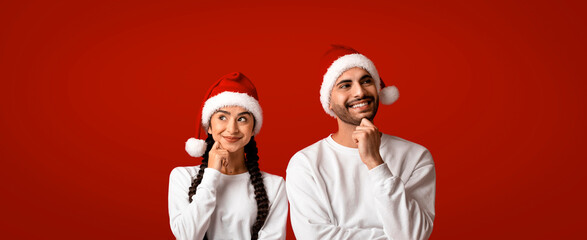 Obraz premium A man and woman are posing thoughtfully while wearing fluffy Santa hats. They are casually dressed in white sweaters and stand in front of a bright red background, capturing a festive mood.