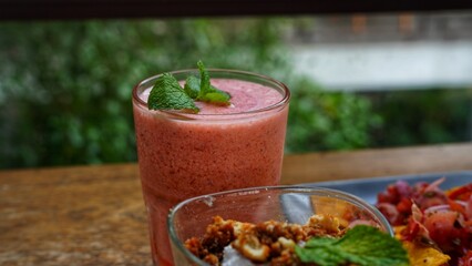 A pink/red beverage, likely a smoothie or juice, served in a tall glass, garnished with mint leaves, against a blurred outdoor background