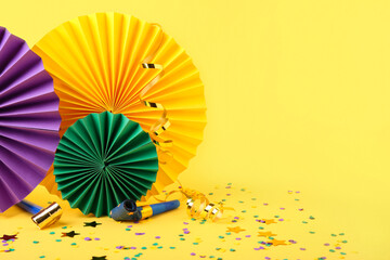 Colorful paper fans with confetti and party blowers on yellow background. Celebration of Mardi Gras