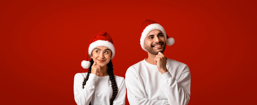 A man and woman are posing thoughtfully while wearing fluffy Santa hats. They are casually dressed in white sweaters and stand in front of a bright red background, capturing a festive mood.