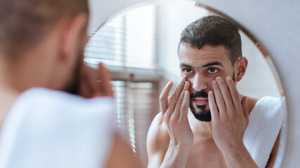 A man stands in front of a bathroom mirror, applying skincare products to his face. The morning...