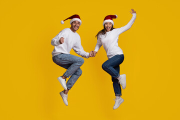 A happy couple jumps in excitement while holding hands, both wearing Santa hats and casual winter outfits. Their bright smiles and vibrant yellow background create a cheerful holiday spirit.