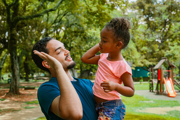 Father and daughter touching their heads playfully while smiling at each other. Warm family connection, natural expressions and joyful childhood lifestyle in a sunny park.