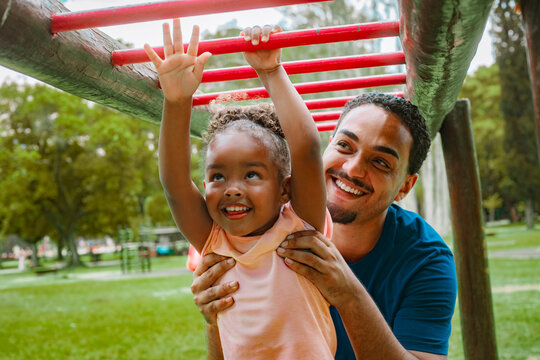 Father helping his daughter on the playground monkey bars, sharing a joyful family moment outdoors. Bright colors, natural expressions and happy childhood vibes in a sunny park setting.