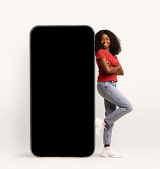 A cheerful Black woman stands confidently while leaning against a big smartphone that has a blank...