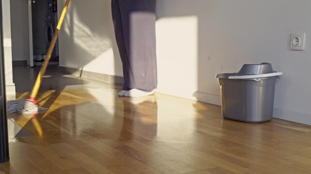 A person diligently mops a shiny wooden floor, showcasing a commitment to a clean and well-maintained home. Sunlight streams across the clean surface.