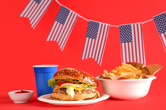 Tasty burger with french fries, flags of USA and cola on red background. American Independence Day celebration