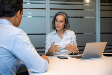 Obraz premium Engaging business meeting in a modern office, featuring a grey-haired woman in a white shirt and a man in a blue shirt, with a laptop and phone on the table.