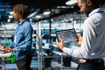 African american data center staff reviews server rigs on industrial platform, using a tablet for data management and innovation. Engineer coordinating infrastructure tasks for machine learning.