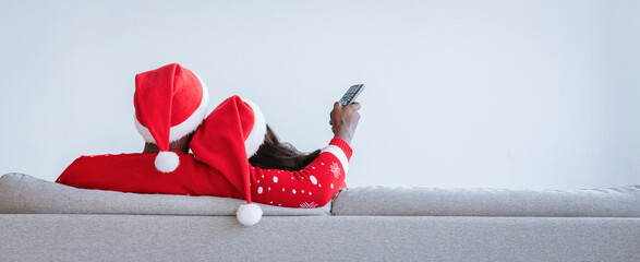 A couple is sitting on a gray sofa, wearing festive Christmas hats, and watching a movie together. They appear relaxed and content, enjoying each other's company in a cozy atmosphere.