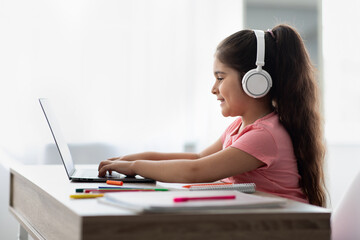 A young girl engages in online learning, focused on her laptop while wearing headphones. She sits at a tidy desk with notebooks and colorful markers nearby, smiling.
