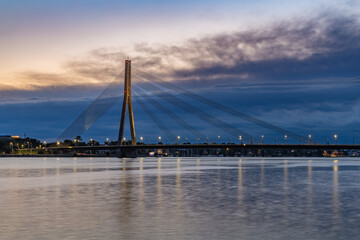 The illuminated Vansu Bridge, a cable-stayed bridge, spans the Daugava River at twilight, with its reflection visible on the calm water and a dramatic cloudy sky overhead