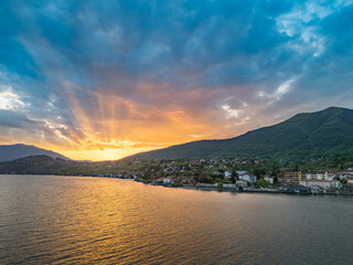 Aerial view of the coastal town of Porto Valtravaglia on Lake Maggiore at sunrise, showing the harbor, colorful buildings, and steep forested hills behind the town