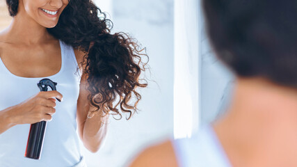 A woman with curly hair is applying hair product from a spray bottle while looking at herself in...