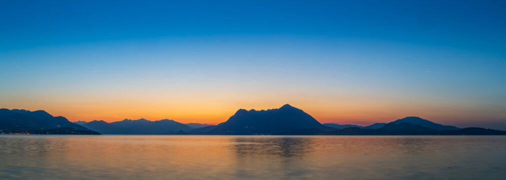 Panoramic view of Lake Maggiore and the silhouette of the surrounding Alps and mountains (likely Mottarone), reflecting the vibrant orange and blue colors of dawn or dusk from Stresa