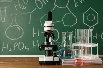 Modern microscope, notebooks and flasks on wooden table near chalkboard with chemical formulas
