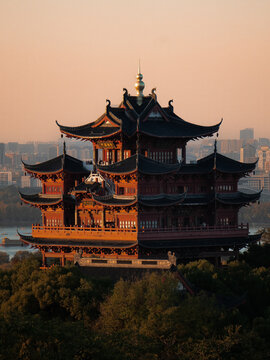 View of a multi-tiered pagoda with dark, ornate roofs stands proudly amidst lush greenery, catching the warm glow of the setting sun, Hangzhou, Zhejiang, China.
