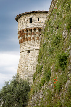 A close-up view of the rugged stone walls and the imposing cylindrical Mirabella Tower of Castello di Brescia, with patches of green vegetation growing on the exterior medieval defenses