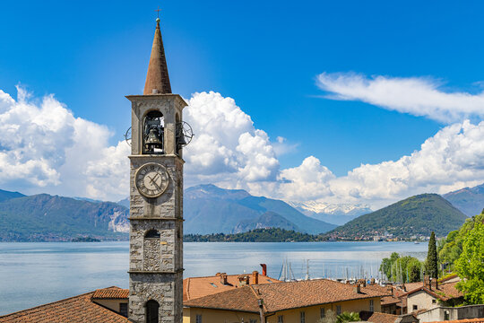 View over the rooftops of Laveno Mombello, featuring the tall stone bell tower and Sant'Ambrogio Church facade, with the blue waters of Lake Maggiore and distant mountains under a cloudy sky
