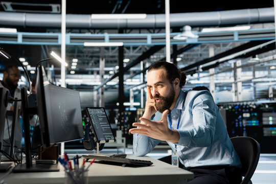 Upset data center engineer monitoring system performance using PC, angry after identifying bottlenecks. Annoyed server farm worker troubleshooting glitches, feeling irked