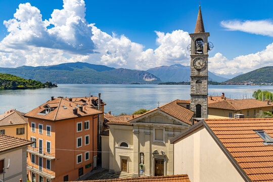 View over the rooftops of Laveno Mombello, featuring the tall stone bell tower and Sant'Ambrogio Church facade, with the blue waters of Lake Maggiore and distant mountains under a cloudy sky