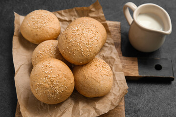 Wooden board of fresh buns with sesame seeds and jug of milk on dark background
