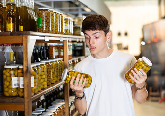 Young guy in supermarket selects examines compares two cans of olives. Male customer chooses jar of...