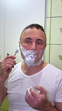 white man shaving with brush and shaving foam looking in the mirror. A man shaves his face in a bathroom with butterflies on the wall and light green tiles.