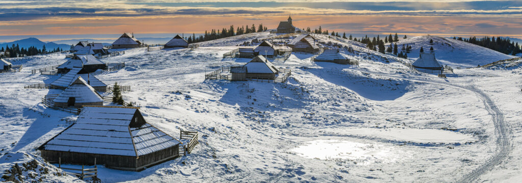 Panoramic view of the snow-covered Velika Planina plateau, showing the traditional wooden shepherd huts and the Chapel of Snow Mary on the hilltop at sunrise