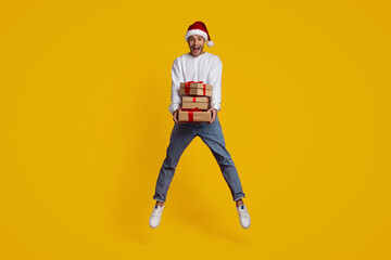 A joyful man in a Santa hat leaps with excitement while holding a stack of wrapped Christmas presents. The bright yellow background enhances the festive mood, showcasing delight and holiday spirit.