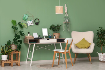 Stylish workplace with tablet on stand, books and different houseplants near green wall at home