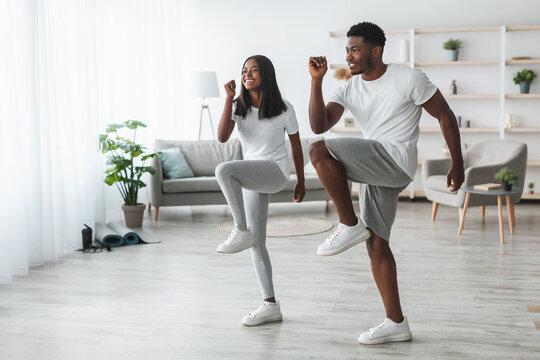 Family Workout. African American Husband And Wife Training Together In Living Room, Doing High Knees Exercise. Happy Black Couple Warming Up, Standing And Lifting Leg Up To Chest, Free Copy Space