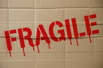 Red, dripping "FRAGILE" warning text stenciled on a corrugated cardboard box, signifying delicate contents requiring careful handling during shipping or storage.